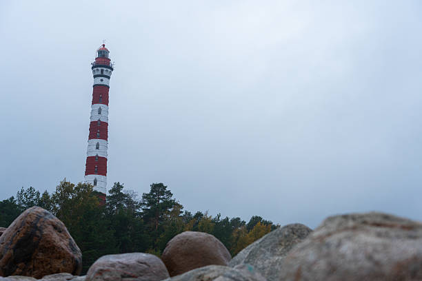 A tall, striped red and white lighthouse rising above a treeline, viewed from behind large coastal boulders on a moody, overcast day