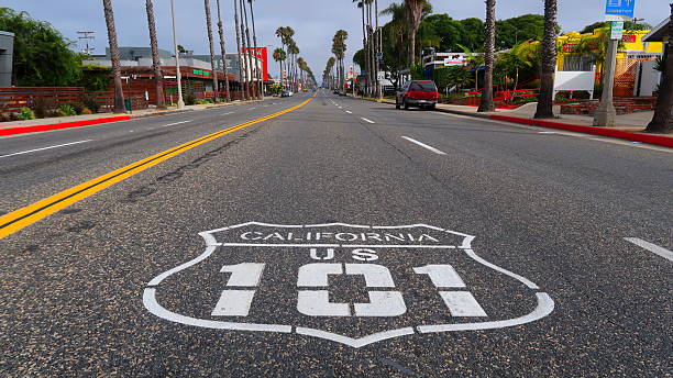 View looking down Coast Highway along U.S. Route 101 in Oceanside, California. The road features the classic “California US 101” shield painted on the asphalt, lined with palm trees, shops, and coastal architecture. This image captures the laid-back Southern California vibe and the charm of a historic Pacific Coast route.