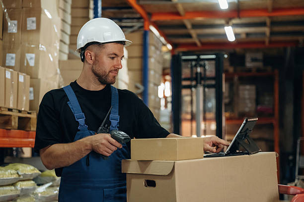 istock In white hard hat, scanning the box. Man in uniform is working in the storage with packages for shipment 2241648895