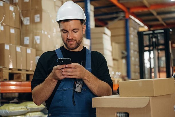 istock Uses smartphone, holding it in hands. Man in uniform is working in the storage with packages for shipment 2241648963