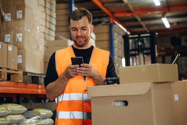 istock Time for a break, using smartphone. Man in uniform is working in the storage with packages for shipment 2241648964