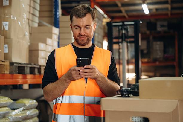 istock Time for a break, using smartphone. Man in uniform is working in the storage with packages for shipment 2241648982