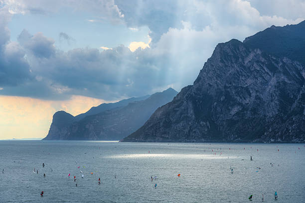Lake Garda panoramic view from Torbole with windsurfers and kitesurfers on water. Dramatic mountains and cloudy sky over the lake, Trentino region, Italy