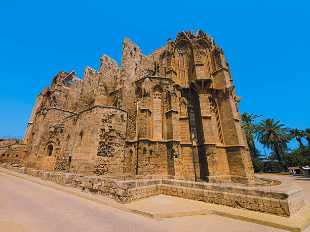 Editorial photo taken on 13 June 2025 in Famagusta, Cyprus, showing the full corner view of the Gothic Saint Nicholas Cathedral, now known as Lala Mustafa Pasha Mosque. The historic structure with pointed arches and flying buttresses is captured under bright sunlight and a vivid blue sky, surrounded by palm trees.