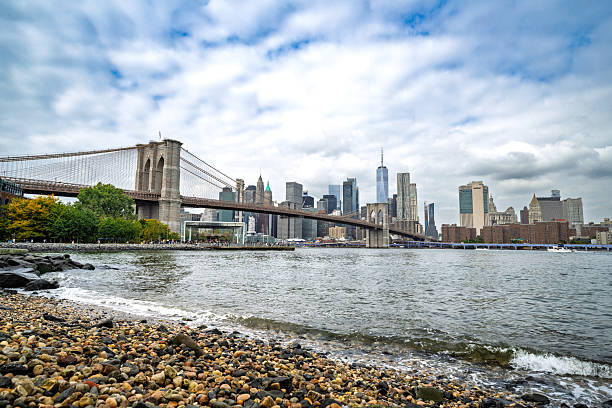 Wide view of the Brooklyn Bridge and the Manhattan skyline seen from Pebble Beach in Dumbo, Brooklyn, New York City. The East River gently reaches the rocky shore under a bright, partly cloudy sky, highlighting one of New York’s most iconic urban landscapes.