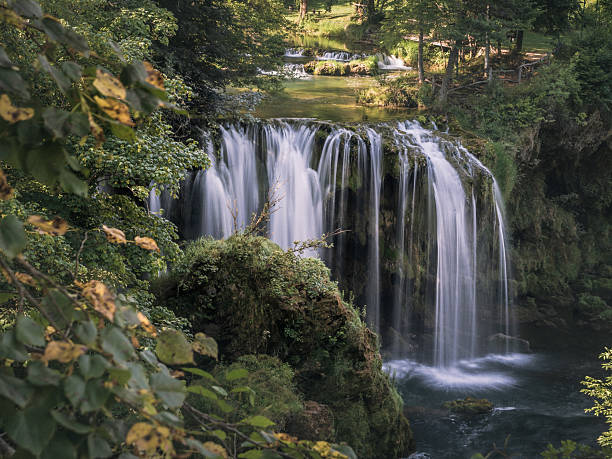 Multi-tiered waterfall pours over mossy rocks into a dark pool, surrounded by dense greenery in the peaceful forest of Rastoke, Croatia.