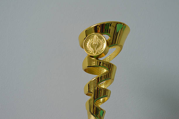 A close-up photo of a golden award trophy featuring a spiral design and a circular emblem with a torch surrounded by laurels. The trophy symbolizes achievement, excellence, and recognition in various competitions or ceremonies