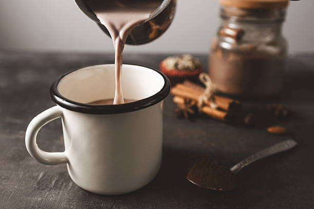 Pouring hot chocolate into white enamel mug on dark rustic surface with cocoa powder, cinnamon sticks, nuts and cupcake, preparing hot chocolate, seasonal cozy winter drink concept.