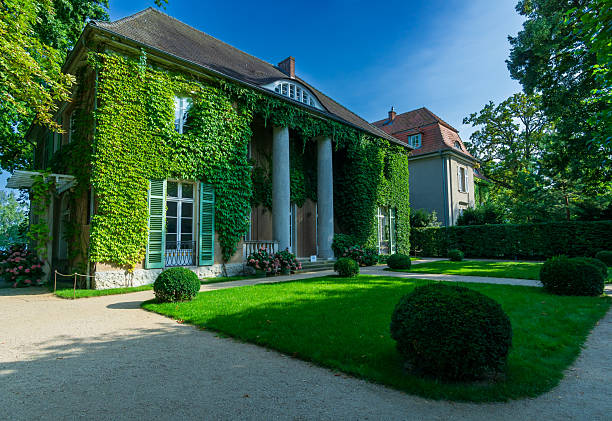 Berlin, Germany - August 9, 2025: Editorial stock photo of the Liebermann Villa in Berlin-Wannsee, Germany. The elegant white villa, once the residence of painter Max Liebermann, is seen from a slightly angled perspective surrounded by lush green gardens and bright summer foliage. The facade is covered in ivy, bathed in warm sunlight under a clear blue sky.