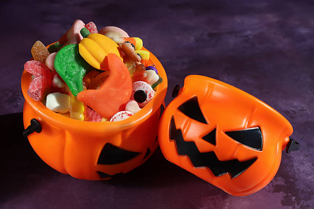 istock Image of two plastic orange Jack O'Lantern pumpkin sweet collection buckets, handled trick or treat containers full of gummy sweets, sweet and sour jellied sweets, mottled purple background, elevated view 2241699622