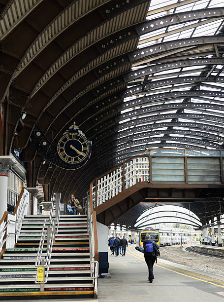 Part of York Railway Station (1877) featuring the station clock and stairs up to a bridge over railway lines. Some passengers are visible and a train is coming in to stop.
