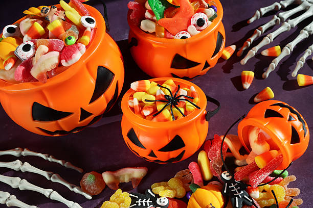 istock Close-up image four plastic, orange Jack O'Lantern pumpkin sweet collection buckets, trick or treat containers full of gummy sweets, surrounded by spilled sweet and sour jellied candy, candy corn, skeletal hands, spider, purple background, elevated view 2241709186