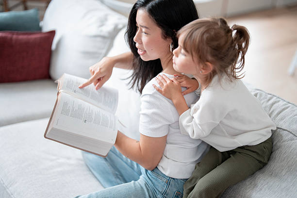 istock Asian Mother Reading Book On Couch While Daughter Hugs Her Back, Encouraging Learning And Bonding 2241709190