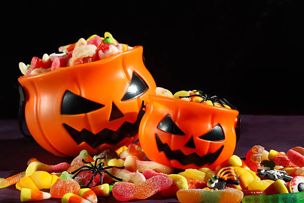istock Close-up image of two plastic, orange Jack O'Lantern pumpkin sweet collection buckets, trick or treat containers full of gummy sweets, surrounded by spilled sweet and sour jellied candy, candy corn, spider, purple and black background, focus on foreground 2241709621
