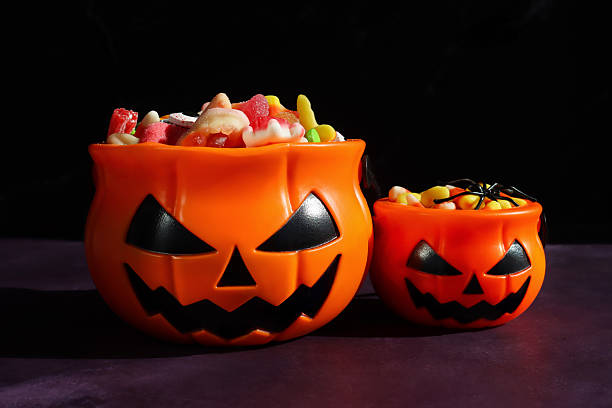 istock Close-up image of two plastic orange Jack O'Lantern pumpkin sweet collection buckets, handled trick or treat containers full of gummy sweets, sweet and sour jellied candy, candy corn, mottled purple and black background, focus on foreground 2241709707