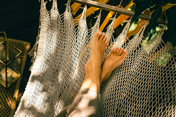 Bare feet rest in a woven hammock outdoors on a warm sunny day with dappled shade, capturing a calm relaxed summer mood in a natural cozy setting