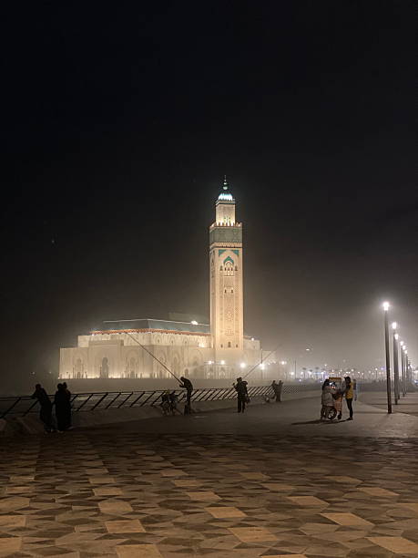 The majestic Hassan II Mosque in Casablanca glowing through evening mist, with its towering minaret reflecting soft light over the Atlantic promenade, as locals stroll and fish by the waterfront.