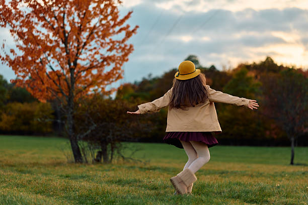 Young girl with hat and coat freely dancing in a green field with fall foliage in the background, experiencing happiness and carefree movement during the golden hour