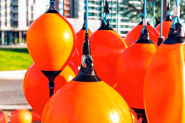 Group of bright orange buoys hang in playground, with modern buildings and city background. Mooring fenders hanging vertically, tied with synthetic lines against brightly urban backdrop