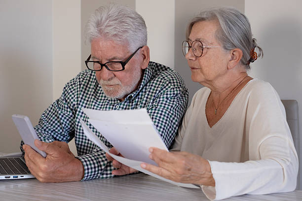 istock Senior couple sitting at home table with paper documents using laptop. Elderly man and woman reading documents, paying bills, managing bank finances, calculating taxes, planning loan, debt, insurance, pension 2241773447