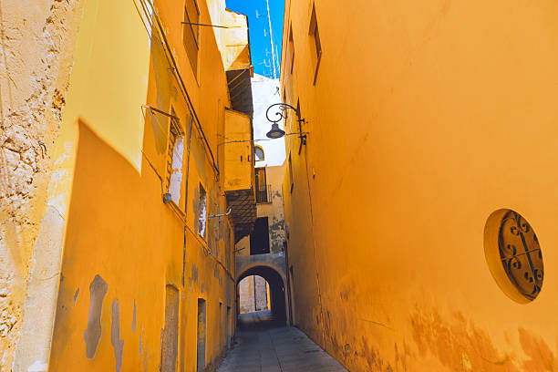 Narrow alleyway winds through yellow buildings with peeling plaster, arched doorways and lanterns casting long shadows. Sky peeks above, framing the rustic Mediterranean charm of weathered stone