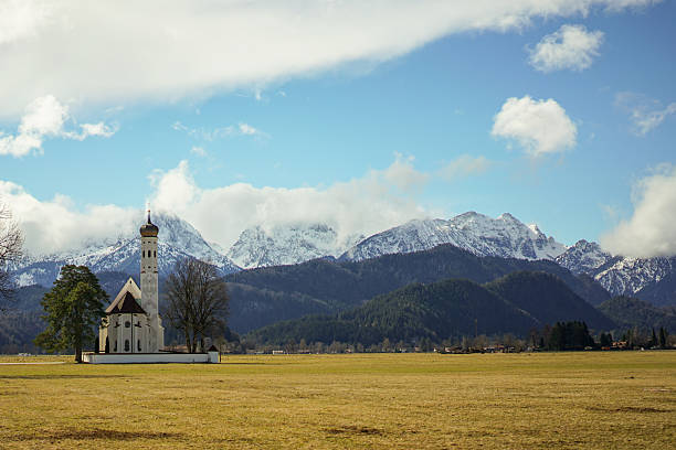 A small white church sits in a field next to a Alps mountain range. The sky is clear and the sun is shining, creating a peaceful and serene atmosphere