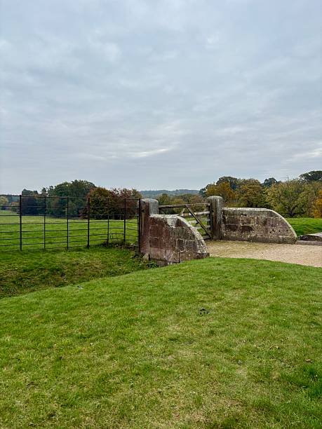 istock A rustic countryside scene featuring a wooden farm gate, moss-covered stone stile, and green pasture with autumn trees in the background. Peaceful rural landscape on an overcast day. 2241803648