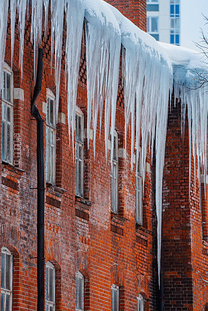 Sharp icicles are hanging from the snowy roof of an old brick building with arched windows, creating a picturesque but dangerous winter scene