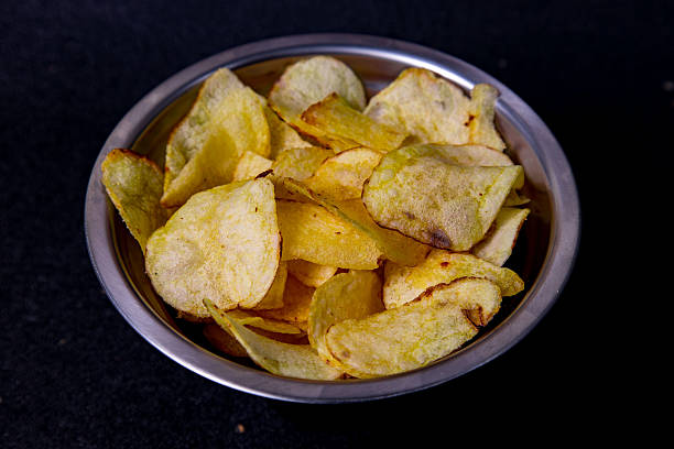 Crunchy potato chips are neatly arranged in a stainless steel bowl set against a dark background, perfect for sharing at a gathering or enjoying while watching a film.