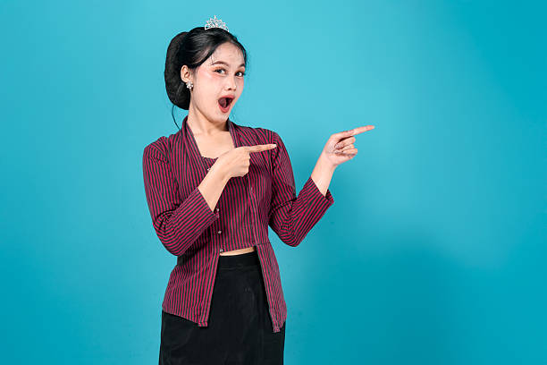istock Charming young Asian woman smiling confidently while pointing to the side, wearing a crown and stylish outfit against blue background, perfect for promotional or advertising content. 2241837792