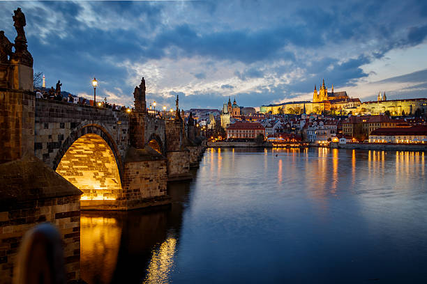 Evening blue hour view of Charles Bridge with glowing lamps, statues, Prague Castle and reflections on the Vltava River
