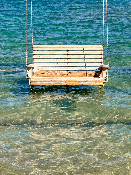 A wooden swing stands partially submerged in shallow seawater near the shore. Gentle waves move beneath it, creating a calm and slightly surreal coastal scene that blends human-made structure with the natural rhythm of the sea.