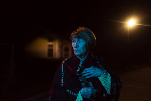 A middle-aged woman stands outdoors at night wrapped in a blanket, showing concern and contemplation about cognitive health. The low light and solitary setting emphasize her emotional state and introspection