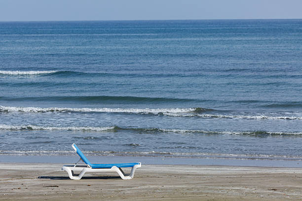 A single, empty white and blue beach lounger rests on a wide stretch of sandy shore. The empty lounger symbolizes a peaceful invitation to relaxation and off-season solitude.