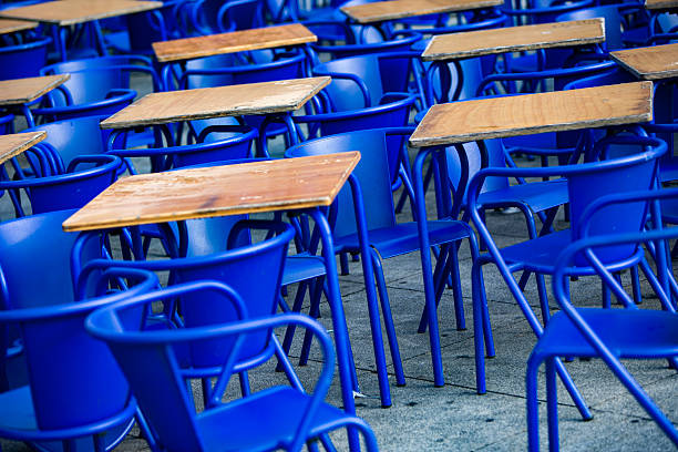Rows of empty blue school-style desks and chairs are arranged outdoors for a public event. A concept of open-air education, community assembly, or a silent, artistic protest.