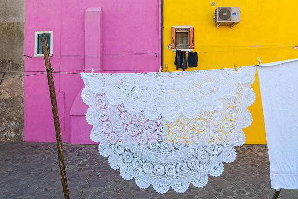 A large, intricately crocheted white tablecloth hangs on a clothesline, drying against vibrant pink and yellow house walls. A concept of traditional craftsmanship and daily life in a colorful village.