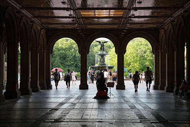 New York City, New York, USA - 07.31.2024: A silhouette of a street musician playing under the arches of Bethesda Terrace frames the famous Angel of the Waters fountain. Tourists and parkgoers enjoy the view and atmosphere on a sunny summer day in New York City.