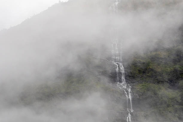 Fog covers mountain with a small, thin waterfall falling down rock face with trees, rocks and bushes near Gudvangen
