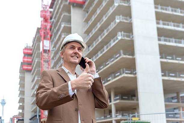Architect in helmet giving thumbs up while on phone at construction site outdoors. stock photo