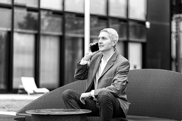 Businessman in blazer talking on phone while seated outdoors on modern chair. stock photo