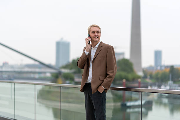 Businessman talking on phone near city riverwalk with modern buildings in background. stock photo