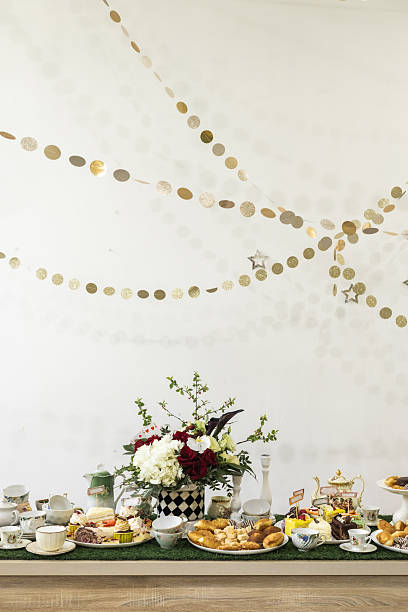 A beautifully decorated tea party table with assorted desserts, floral centerpiece, and festive golden garlands against a clean white wall background.