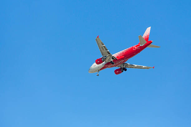 Underside view of a Russian Rossiya Airlines aircraft operated by Russia, captured mid-flight against a clear blue sky. The plane features a red and white fuselage with a pattern of red shapes on the tail and rear section, and the airline name “РОССИЯ” prominently displayed. The extended landing gear and dynamic angle highlight the aircraft’s motion and national branding, ideal for aviation visuals and Eastern European travel themes.