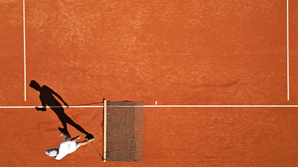 Tennis court maintenance crew cleans surface on a clay court, captured mid-action with a dynamic shadow. The scene highlights sports training and skill development, emphasizing the textured clay surface and focused athletic movement. Ideal for tennis education and training class imagery., captured with a top-down view.