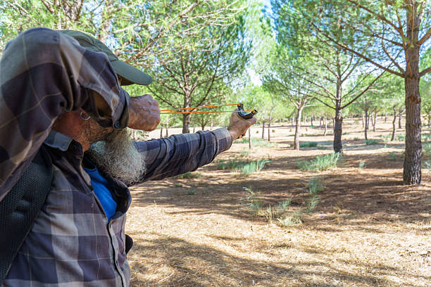 Man with beard and plaid shirt aiming a slingshot, focusing intently on a target in a pine forest setting