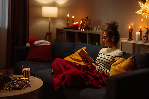 Cozy Christmas night: woman reading book on sofa with warm lights, blankets and candles