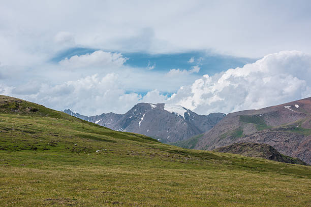 istock Dramatic alpine view from green hills to large rocky mountain range with glacier under clouds in blue sky. Scenic landscape with big snow mountain top in center. Changeable weather in high mountains. 2241931741