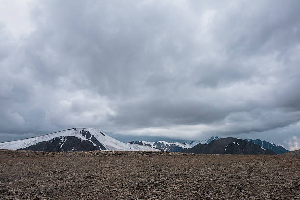 istock Dramatic misty top view from stone pass to big glacier tongue among sharp rocks and large snow-capped mountains in rainy low clouds. Dark atmospheric mountain silhouettes in rain in gray cloudy sky. 2241932628