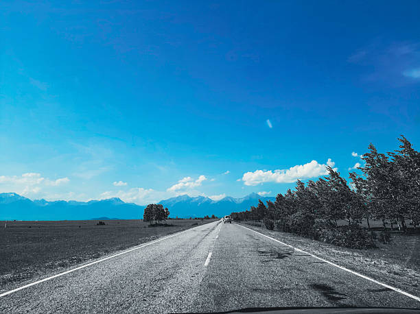 A view from a car onto a small country road with smooth asphalt, surrounded by fields, forests, and mountains on the horizon. Cinematic blue-graphit depth effect.