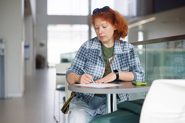 Gen X woman with red curly hair writes notes with pen on paper at round table in community center workspace. She wears sunglasses on head and smartwatch while working. Laptop sits open on table beside her. Natural daylight fills the modern interior space through large windows. Selective focus on woman with blurred background. Medium shot captures upper body and workspace.
